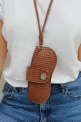 Cuelga lentes de cuero color chocolate con textura trenzada, diseñado con un estuche protector integrado que se cierra mediante un broche metálico a presión. Incluye una correa larga para llevar colgado al cuello.