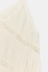 Top asimétrico de viscosa en color crudo, con escote en V y tirantes finos ajustables con lazadas. Presenta pliegues y detalles de entredós de encaje en diagonal, terminando en un bajo asimétrico con volados.