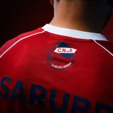 Camiseta de fútbol Umbro del Club Nacional de Football, modelo Away 2026. Diseño en color rojo con un patrón texturizado de ADN en relieve, cuello en V con detalles en azul marino y blanco. Incluye logo de Umbro, escudo del club con las tres copas intercontinentales bordadas, y sponsors estampados en el frente y mangas.