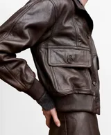 Chaqueta bomber de cuero ovino color marrón oscuro, con cierre frontal de botones, bolsillos con solapa en el pecho y cuello alto con broches.