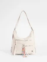 Mochila tipo cartera hobo color beige, con diseño versátil que permite llevarla de ambas formas. Cuenta con múltiples bolsillos frontales con cierre, tiradores largos y un llavero decorativo removible con cuentas coloridas. Incluye correa regulable y herrajes metálicos plateados.