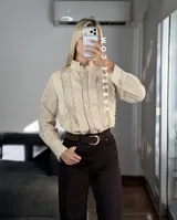 Camisa blanca de estilo romántico con cuello alto, cierre frontal de botones y detalles de encaje y alforzas verticales. Presenta mangas largas con puños decorados con encaje.