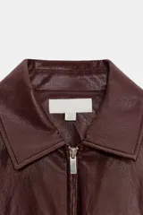 Chaqueta efecto piel color marrón, con cuello solapa, mangas por debajo del codo, cintura elástica, falsos bolsillos delanteros y cierre frontal con cremallera.
