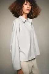 Camisa de popelín color blanco, con cuello solapa, escote en pico y cierre frontal con botones. Presenta un corte holgado y mangas largas con puños rectos.
