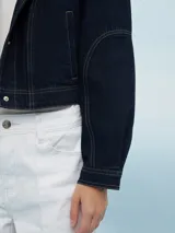 Campera de jean azul claro, estilo trucker, con cuello mao y botones metálicos plateados. Presenta costuras decorativas en el frente y mangas largas con puños doblados.