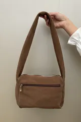 Cartera de hombro confeccionada en gamuza color habano, con diseño de correa ancha decorada con tachas metálicas circulares. Presenta cierre superior y placa metálica con logo en el frente.
