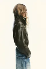 Cazadora corta de efecto piel color marrón oscuro, con cuello subido, manga larga con trabillas en los hombros, bolsillos con solapa en el delantero y cierre frontal con cremallera y botones a presión ocultos por solapa.