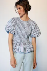 Top de cuello redondo con cuerpo de nido de abeja elástico y mangas globo con elástico en los puños. Presenta un diseño a rayas verticales en tonos azul, blanco y granate, con un bajo estilo peplum.