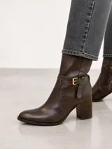 Bota de caña media confeccionada en cuero color marrón oscuro, con punta almendrada y taco cuadrado de 7 cm. Presenta una tira decorativa con hebilla metálica dorada en la zona del tobillo.