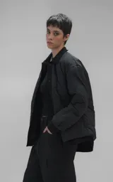 Campera tipo sobrecamisa de diseño oversize, confeccionada en microfibra opaca con patrón matelaseado geométrico. Presenta cuello, puños, cartera central y ruedo en gabardina de algodón, con cierre frontal mediante broches a presión. El diseño del ruedo simula una prenda superpuesta.