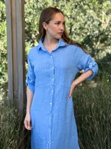 Vestido camisero largo de lino blanco, con cuello camisero, manga corta enrollada y cierre frontal de botones de nácar. Presenta aberturas laterales en el bajo.