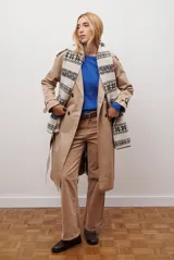 Trench coat color beige con cuello de solapa, cierre frontal abotonado, bolsillos laterales y mangas largas con cintas ajustables. Incluye capucha desmontable y forro interior rayado. La tela exterior cuenta con protección repelente a la lluvia y manchas.