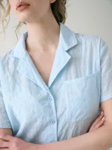 Camisa de manga corta para dormir confeccionada en broderie de algodón, con cuello solapa y cierre frontal con botones. Presenta un bolsillo aplicado en el pecho.