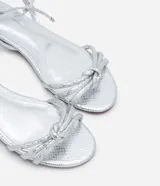 Sandalia plana marca Satinato, color dorado metalizado con textura tipo croco. Posee tiras finas con detalle de nudo en el empeine y cierre de lazo ajustable al tobillo. Diseño de puntera redonda y suela plana.