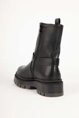Bota estilo biker de cuero sintético color negro, con suela tipo plataforma dentada. Presenta una tira decorativa con argolla metálica y apliques de tachas en el tobillo.