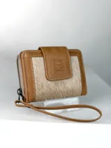 Billetera pequeña de cuero color marrón claro con paneles frontales de pelo natural. Presenta un cierre de solapa con broche y un logo grabado de un búho. Incluye una correa de muñeca extraíble.