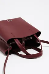 Bolso pequeño de piel color borgoña, con estructura rígida y forma de cubo. Posee doble asa de mano y una correa larga para llevar al hombro o cruzado. El frente lleva el logo metálico de Bimba y Lola.