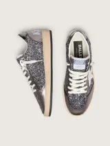 Zapatillas Golden Goose modelo Ball Star de estilo retro, con empeine y lengüeta cubiertos de glitter gris oscuro y detalles en cuero laminado. Presentan la clásica estrella lateral en cuero blanco, puntera reforzada y cordones de algodón crudo, con suela de goma desgastada.