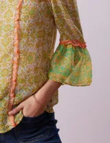 Blusa de gasa estampada con flores amarillas y verdes, marca Alex & Lili. Tiene cuello mao, mangas 3/4 acampanadas y cierre frontal con botones. Presenta volados verticales color naranja en el frente y en las mangas.