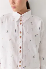 Camisa de lino blanca con estampado de figuras deportivas pequeñas y detalles de costuras en color rojo. Presenta cuello clásico, cierre frontal con botones de madera, mangas largas con puños abotonados y un bolsillo frontal aplicado.