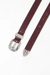 Cinturón de cuero con textura efecto cocodrilo en color bordó, con hebilla y pasador metálicos plateados de estilo western con detalles grabados.