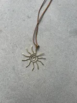 Collar con dije de bronce en forma de sol con diseño de espiral y rayos ondulados, colgando de un tiento de gamuza marrón.