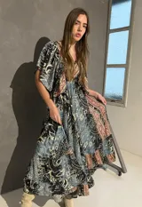 Vestido largo con escote en V tanto en el frente como en la espalda, mangas cortas amplias y corte elastizado debajo del busto. Presenta un estampado de estilo paisley y guardas geométricas en tonos azules, negros y corales.