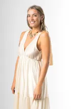 Vestido largo color crudo, confeccionado en mezcla de tencel y lino, con escote en V pronunciado, breteles anchos terminados en lazos en los hombros, y falda larga de varios niveles con volados amplios y fluidos.