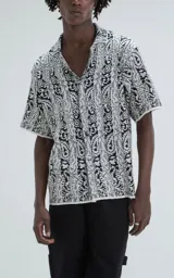 Camisa de manga corta con tejido de punto abierto tipo crochet, corte oversize y cuello solapa. Presenta un diseño estampado con rayas verticales y motivos paisley en blanco y negro.