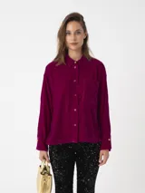 Camisa de terciopelo color fucsia, de corte recto y holgado, con cuello camisero, cierre frontal con botones y bolsillo de parche en el pecho.