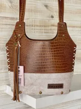 Bolso matero de cuero marrón con textura tipo cocodrilo en la parte superior y cuero liso color beige en la parte inferior. Tiene doble asa de cuero, costuras blancas a la vista y detalles de tachas metálicas. Incluye colgante de cuero y forro interior.