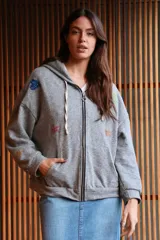 Campera con capucha y cierre frontal, confeccionada en algodón gris jaspeado. Presenta detalles decorativos de figuras como corazones, estrellas y coronas con acabado brillante en diversos colores. Cuenta con bolsillos laterales y cordones ajustables en la capucha.