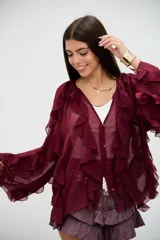 Blusa bordó de tela semitransparente con diseño de volados verticales en el frente y mangas largas acampanadas. Presenta cierre frontal con botones y detalles de bordados circulares sutiles en la parte inferior.