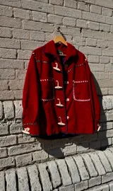 Chaqueta roja de cuero 100% artesanal, con pasamanería y detalles de costura en tiento. Posee bolsillos plaqué frontales, cuello camisero, botones de cuero alargados tipo duffel y forro interno.