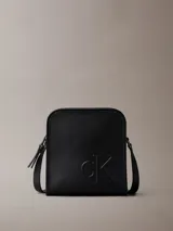 Cartera tipo reporter de Calvin Klein confeccionada en piel sintética suave de color negro. Presenta un diseño cuadrado con monograma tonal grabado en el frente, correa ajustable para cruzar, cierre de cremallera superior y bolsillo exterior trasero.