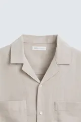 Camisa de lino de calce relajado en color beige, con cuello bowling y manga corta. Cuenta con dos bolsillos de parche en el pecho, aberturas laterales en el bajo y cierre frontal con botones.