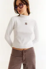 Top de manga larga con cuello alto, confeccionado en tejido de lycra y algodón. Presenta costuras a contratono, bordado central en el pecho, aberturas laterales en el ruedo y diseño con dedal en las mangas.