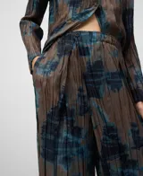 Pantalón ancho de tiro alto con efecto plisado (crinkle) y estampado abstracto en tonos marrón topo y azul. Presenta un diseño cruzado en el frente con cierre de botón y cintura elástica.
