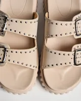 Sandalias tipo mule de color beige, con suela de plataforma texturizada. Posee dos tiras anchas en el empeine con hebillas metálicas cuadradas y decoración de remaches plateados.
