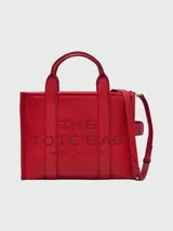 Bolso tipo tote de cuero negro, modelo The Leather Medium Tote Bag de Marc Jacobs. Presenta doble asa superior, correa ajustable para el hombro y el texto grabado "THE TOTE BAG MARC JACOBS" en el frente.