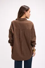 Camisa de corte oversize con estampado de rayas verticales finas en color marrón sobre fondo blanco. Presenta cuello clásico, cierre frontal con botones, bolsillo en el pecho y mangas largas con puños abotonados.