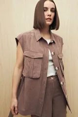 Chaleco color beige con cuello camisero, sin mangas, cierre frontal con botones y cinturón de lazo. Presenta bolsillos de parche en el pecho y pinzas verticales en el frente.