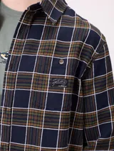 Camisa de manga larga con estampado de cuadros en tonos blanco, gris y azul. Presenta cuello clásico, cierre frontal con botones y un pequeño parche con logo en el pecho.