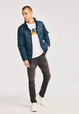 Chaqueta acolchada de peso medio en color azul petróleo, con cierre frontal de cremallera, cuello alto y parche de silicona con el logotipo de la marca en el pecho. Cuenta con bolsillos laterales y ribetes elásticos en puños y dobladillo.