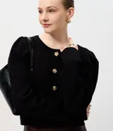 Cardigan de punto negro con cuello redondo, mangas largas abullonadas y cierre frontal con botones decorativos en forma de flor dorada. Presenta un corte corto y ajustado.