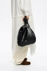 Bolso de mano mediano de piel vacuna, color negro, con estructura blanda y forma de cubo o 'paper bag'. Posee asas de mano cortas y una correa ajustable y extraíble de tela azul marino. El logo de Bimba y Lola está grabado en la parte inferior frontal.