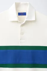 Remera polo de manga larga de corte regular fit, confeccionada en algodón color blanco crudo. Presenta cuello solapa blanco, cierre frontal con botonadura oculta y una franja horizontal ancha en el pecho en color azul y verde.