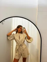 Mono corto de lino color beige, con cuello camisero, cierre frontal, mangas largas abullonadas y cinto ajustable en la cintura.