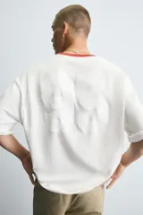 Camiseta de corte relajado, color blanco, con cuello redondo en contraste rojo y manga corta. Presenta un pequeño estampado en el pecho con texto cursivo y el número "99" en color blanco tenue.