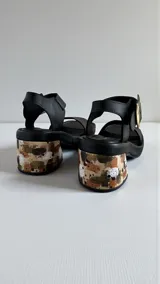 Sandalias de cuero negro Carolina De Cunto, con taco bloque medio intervenido con diseño artístico y hebilla metálica grande en tono oro viejo con logo grabado.